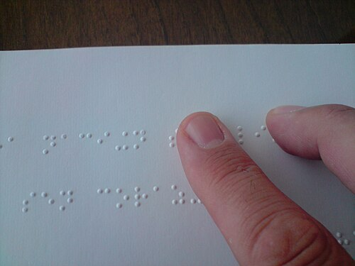 Zeige- und Mittelfinger eines Lesenden gleiten über eine Zeile Braille-Text. Deutlich zu erkennen sind die punktförmigen Erhöhungen im weissen Papier, die die Schriftzeichen bilden.